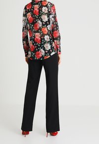 Blusa nera floreale con rose rosse e rosa, tessuto trasparente, maniche lunghe, abbinata a pantaloni neri e tacchi alti rossi.