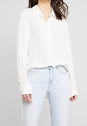 Kobieta ubrana w luźną białą bluzkę z długim rękawem wpuszczoną w jasnoniebieskie, biodrówki, stojąca na prostym tle.