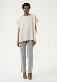 Lichtbeige poncho-stijl top met golvende halslijn, gecombineerd met rechtvallende grijze jeans en tan enkelboots, tegen een witte achtergrond.