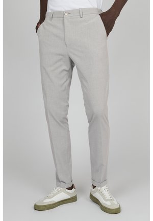 Mężczyzna ubrany w jasnoszare, dopasowane spodnie oraz białe trampki z zielonymi podeszwami, stojący z rękami w kieszeniach na jednolitym tle.