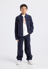 Laivansininen sametinen takki, jossa on kaksi etutaskua, yhdistettynä yhteensopiviin housuihin. Alla valkoinen t-paita, jossa on oranssi graafinen kuva. Valkoiset lenkkarit.