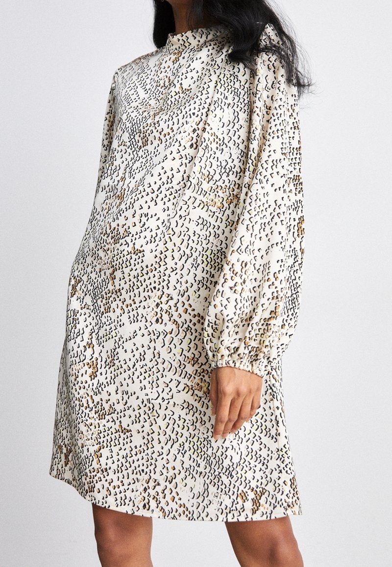 Femme portant une robe ample à manches longues et col montant avec un motif abstrait beige et noir, longueur au-dessus du genou.