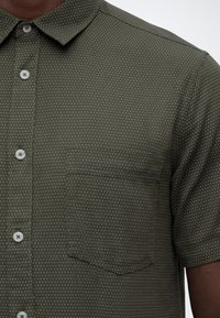 Ανδρικό πουκάμισο με κοντό μανίκι σε ελιάς πράσινο χρώμα, με μικρό μοτίβο λευκών τελειών και τσέπη στο στήθος.