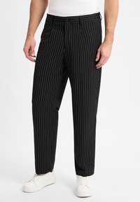 Zwarte pinstriped broek met een getailleerde pasvorm, voorzien van een haak- en ritssluiting, voorplooien, ceintuurlussen en gedragen met witte sneakers.