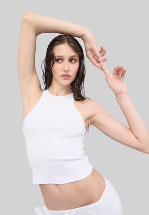 Top de tirantes blanco con canalé y cuello alto, combinado con pantalones claros y holgados. El modelo posa con los brazos levantados, sobre un fondo gris.