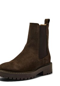 Bota Chelsea de ante marrón con paneles elásticos en los lados, una lengüeta para tirar y una suela de goma texturizada con tacos profundos para agarre y tracción.