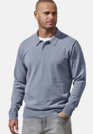 Mann mit kurzen Haaren, der ein hellblaues Polohemd mit langen Ärmeln und graue Jeans trägt, steht mit einer Hand in der Tasche vor einem schlichten Hintergrund.