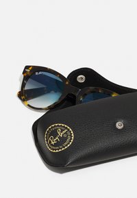Occhiali da sole oversized a forma di occhio di gatto con motivo tartarugato, lenti blu scuro e una custodia in pelle nera con logo dorato e chiusura a scatto.