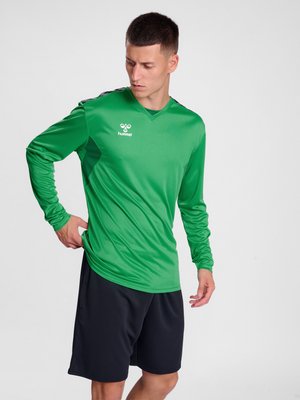 Mand iført en grøn langærmet sports-trøje og sorte shorts, justerer trøjens kant, står mod en ensartet lys baggrund.