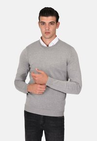 Maglione grigio lavorato a maglia con scollo tondo, maniche lunghe e polsini e orlo a costine, indossato sopra una camicia bianca con colletto.