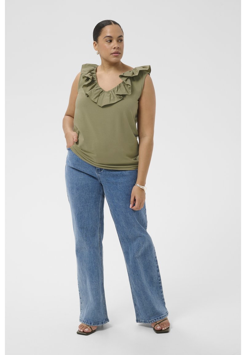 Femme portant un top sans manches vert olive avec un col à volants et un jean bleu à jambes larges, debout avec une main dans la poche.