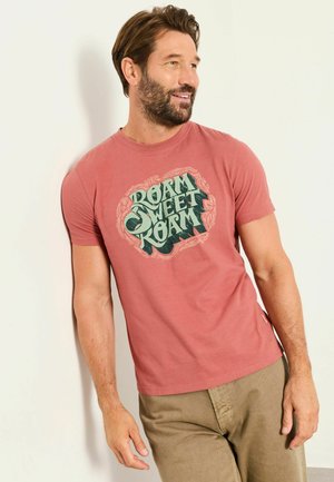 Mann mit Bart trägt ein ausgewaschenes rotes T-Shirt mit dem Text "Roam Sweet Roam", lehnt an einer weißen Wand, lächelt und schaut nach rechts.