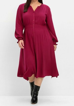 Frau trägt ein langärmliges, knopfgeschlossenes burgunderfarbenes Midikleid mit taillierter Taille und schwarzen Stiefeletten vor einfarbigem Hintergrund.