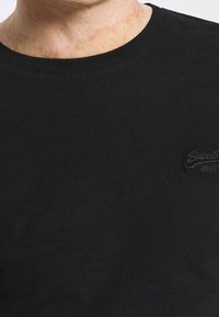 Schwarzes Baumwoll-T-Shirt mit Rundhalsausschnitt, das ein dezentes prägbares Logo in der oberen linken Ecke aufweist. Glatte Oberfläche ohne sichtbare Muster.