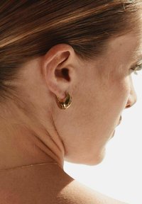 Perfil derecho de mujer mostrando oreja con pendiente de aro dorado, cabello castaño claro recogido hacia atrás y un collar delgado de oro en el cuello.