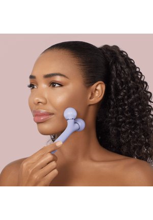 Femme aux cheveux bouclés utilisant un rouleau de massage facial à la lavande sur sa joue sur un fond rose uni.