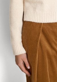 Cremfarbener Strickpullover mit gerippten Bündchen, kombiniert mit einem rostfarbenen Cordrock mit vertikalen Streifen und einem geplätteten Design.
