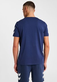 Navyblaues Baumwoll-T-Shirt mit kurzen Ärmeln, das weiße Chevron-Muster an den Schultern aufweist. Klassischer Rundhalsausschnitt, lockere Passform.