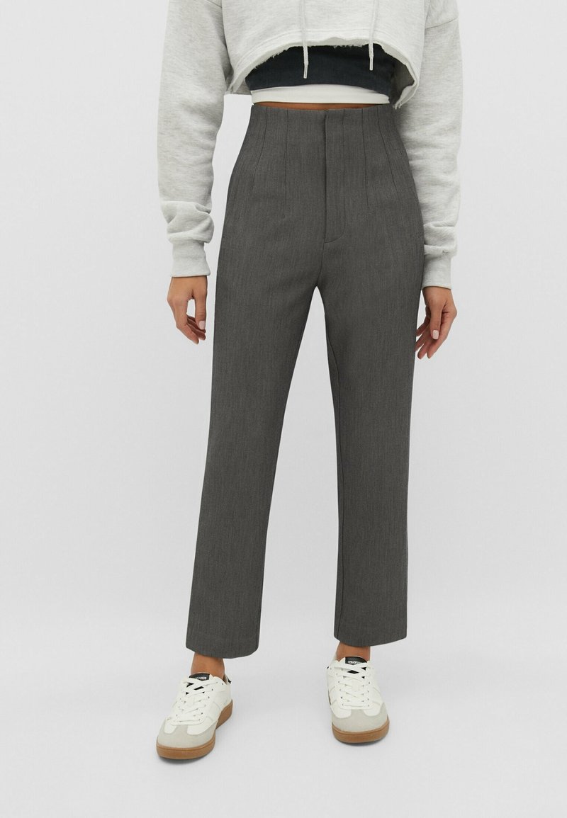 Femme portant un pantalon gris droit taille haute, baskets blanches, sweat à capuche gris court et top noir court sur un fond uni.