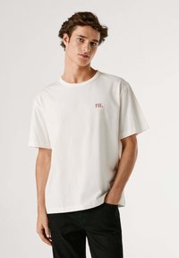 Maglietta bianca in cotone con maniche corte. Presenta un piccolo ricamo rosso "PJL" sul petto a sinistra e una vestibilità comoda. Pantaloni neri visibili.