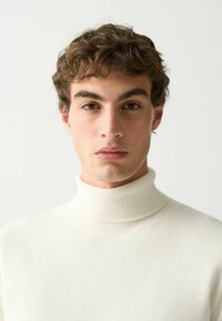 Bijela, rebrasta turtleneck pulover od mekog materijala, sa visokim ovratnikom i prilagođenim dizajnom, nosi osoba sa kratkom kosom.