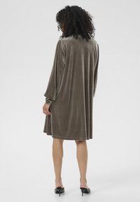 Frau mit lockigem Haar, die ein lockeres, knielanges braunes Samtkleid und schwarze, offene Absatzschuhe trägt, steht mit dem Rücken zu einem schlichten Hintergrund.