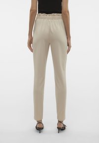 Beige, strakke broek met een geplooide elastische tailleband. Gladde textuur en taps toelopende broekspijpen, gecombineerd met zwarte sleehaksandalen.