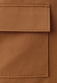 Gros plan sur un tissu marron avec un rabat rectangulaire à double couture et une poche, montrant la texture détaillée et les lignes de couture.