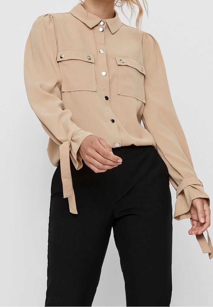 Blouse beige à boutons avec deux poches poitrine et manches longues nouées aux poignets, associée à un pantalon noir taille haute.
