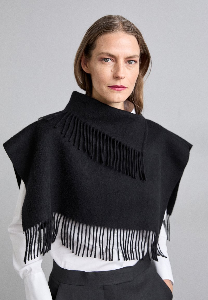Black poncho made of soft wool, featuring a high collar and long fringes along the bottom edge. Accompanied by a white shirt underneath.