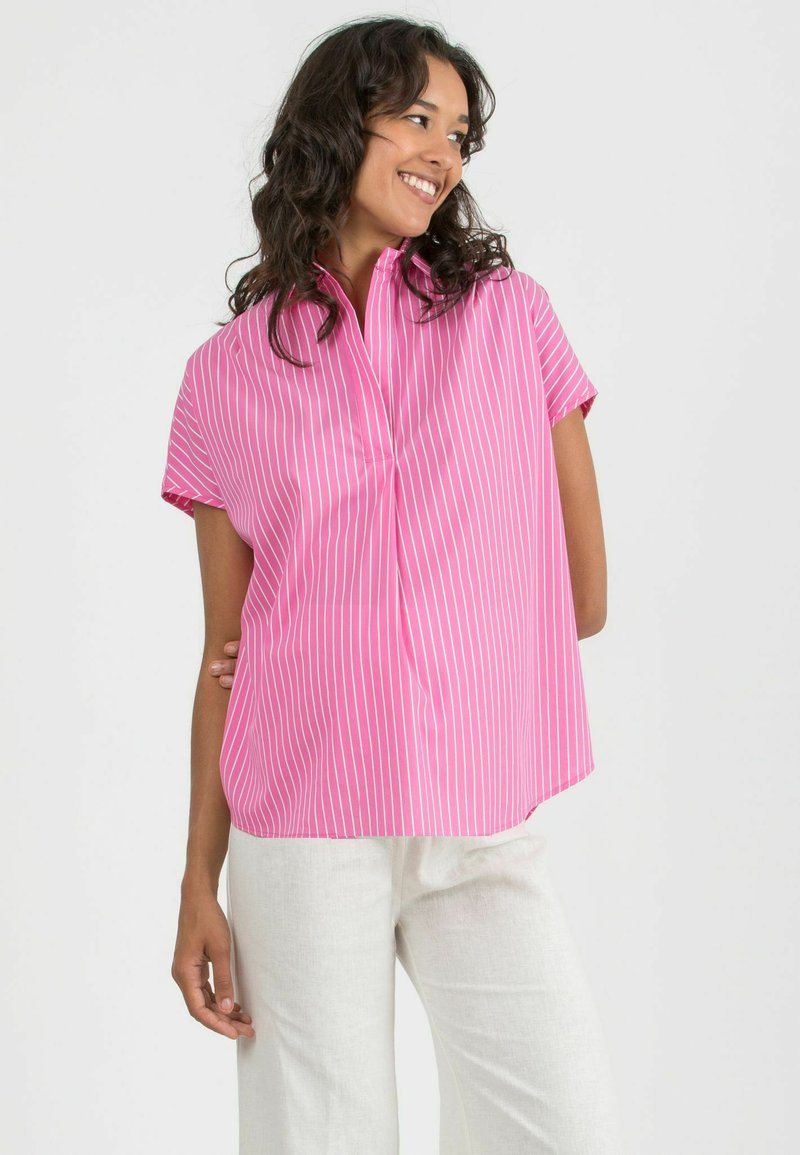 Camicia a maniche corte con colletto in vivace rosa e righe verticali bianche, caratterizzata da una vestibilità comoda e un tessuto morbido, indossata con pantaloni color crema.