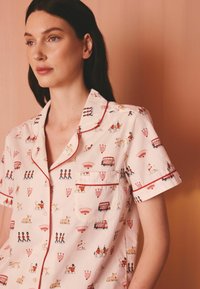Femeie purtând o cămașă roz deschis cu mâneci scurte, cu nasturi și detalii roșii, decorată cu imprimeuri tematice Londra, inclusiv gărzi, autobuze și câini.
