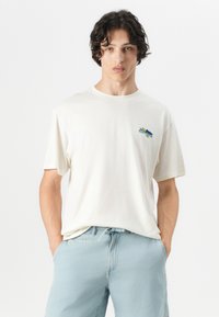 Cream-colored cotton T-shirt with short sleeves, featuring a small colorful logo on the left chest. Paired with light blue shorts.