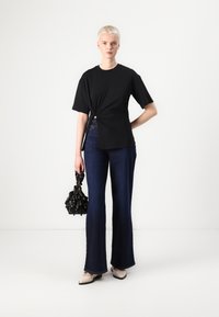 Tricou scurt cu manșete negre și noduri, blugi evazați în nuanță de albastru închis, flats crem și o geantă neagră cu accente florale, prezentate pe un fundal neutru.