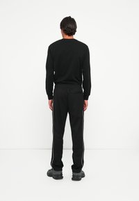 Man in zwart langemouw shirt en zwarte broek met witte zijstreep staat met zijn rug naar de camera tegen een eenvoudige witte achtergrond.