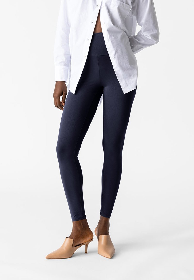 Granatowe legginsy z gładkiego, elastycznego materiału, zestawione z białą koszulą i beżowymi szpilkami, w których zaprojektowano szpiczasty czubek.
