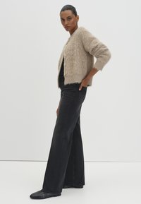 Tan fuzzy cardigan with long sleeves, single-button closure, paired with high-waisted black flared jeans and black shoes. Simple design.