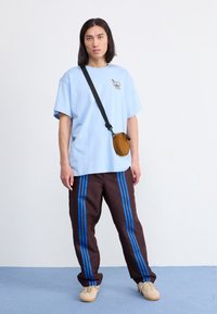 Světlounce modré oversize tričko s logem srdce; hnědé vzorované kalhoty s modrými pruhy. Modelka drží hnědou kabelku přes rameno, má na sobě béžové boty.