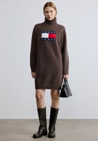 Καφέ πλεκτό φόρεμα με ψηλό λαιμό, με έντονο λογότυπο "TOMMY JEANS". Συνδυασμένο με ψηλές μαύρες δερμάτινες μπότες και μικρή μαύρη τσάντα.