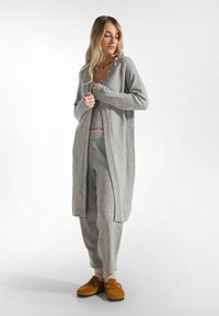 Helle graue, gemütliche Strickjacke über einem passenden Oberteil und einer Hose. Weiche Textur, lässige Passform. Braune Hausschuhe ergänzen die warmen, neutralen Töne.