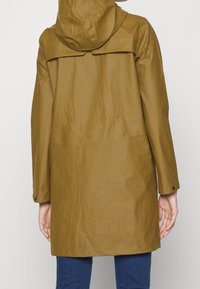 Cappotto impermeabile marrone con cappuccio, dotato di yoke sul retro, maniche lunghe e polsini con bottone a pressione. Texture liscia e design fino al ginocchio.