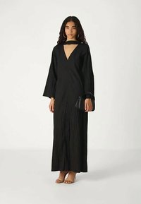 Žena v dlhých čiernych šatách s hlbokým V-výstrihom a širokými rukávmi, drží malú čiernu kabelku a má na sebe béžové podpätky.