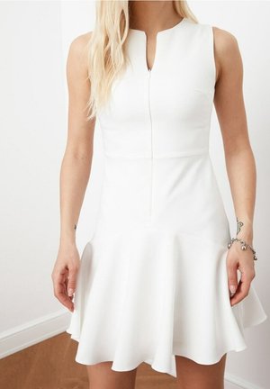 Biała sukienka bez rękawów z zamkiem z przodu, dopasowanym gorsetem i rozkloszowaną spódnicą, noszona przez osobę o długich blond włosach i bransoletkach na nadgarstku.