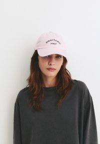 Lichtroze katoenen baseballcap met zwarte geborduurde tekst "POSITANO ITALIË." Model draagt een donkergrijze langemouwen T-shirt.