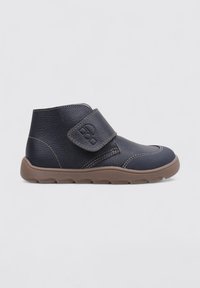 Zapato negro de cuero hasta el tobillo con suela marrón texturizada, cierre de tira de velcro y área de punta reforzada.
