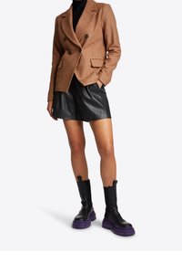 Ruskea houndstooth-mallinen bleiseri kaksoispainikoinnin kera, yhdistettynä mustiin nahkashortseihin ja näyttäviin mustiin saappaisiin, joissa on violetit pohjat.