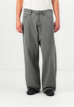 Mężczyzna ubrany w luźne, wyblakłe szare dżinsy, czarne buty i białą koszulę, stojący na tle jednolitego białego tła.