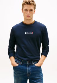 Camicia a maniche lunghe blu navy con logo "TOMMY HILFIGER" in bianco, rosso e blu; abbinata a jeans di denim blu. Tessuto liscio, vestibilità comoda.
