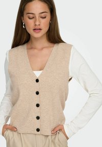Un gilet di maglia senza maniche beige con scollatura a V profonda e sei bottoni neri lungo la parte anteriore. Abbinato a una camicia bianca a maniche lunghe sotto.
