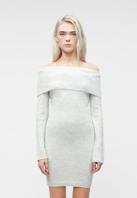 Kvinne med langt blondt hår iført en lys grå avskåret strikket kjole med lange ermer mot en enkel hvit bakgrunn.